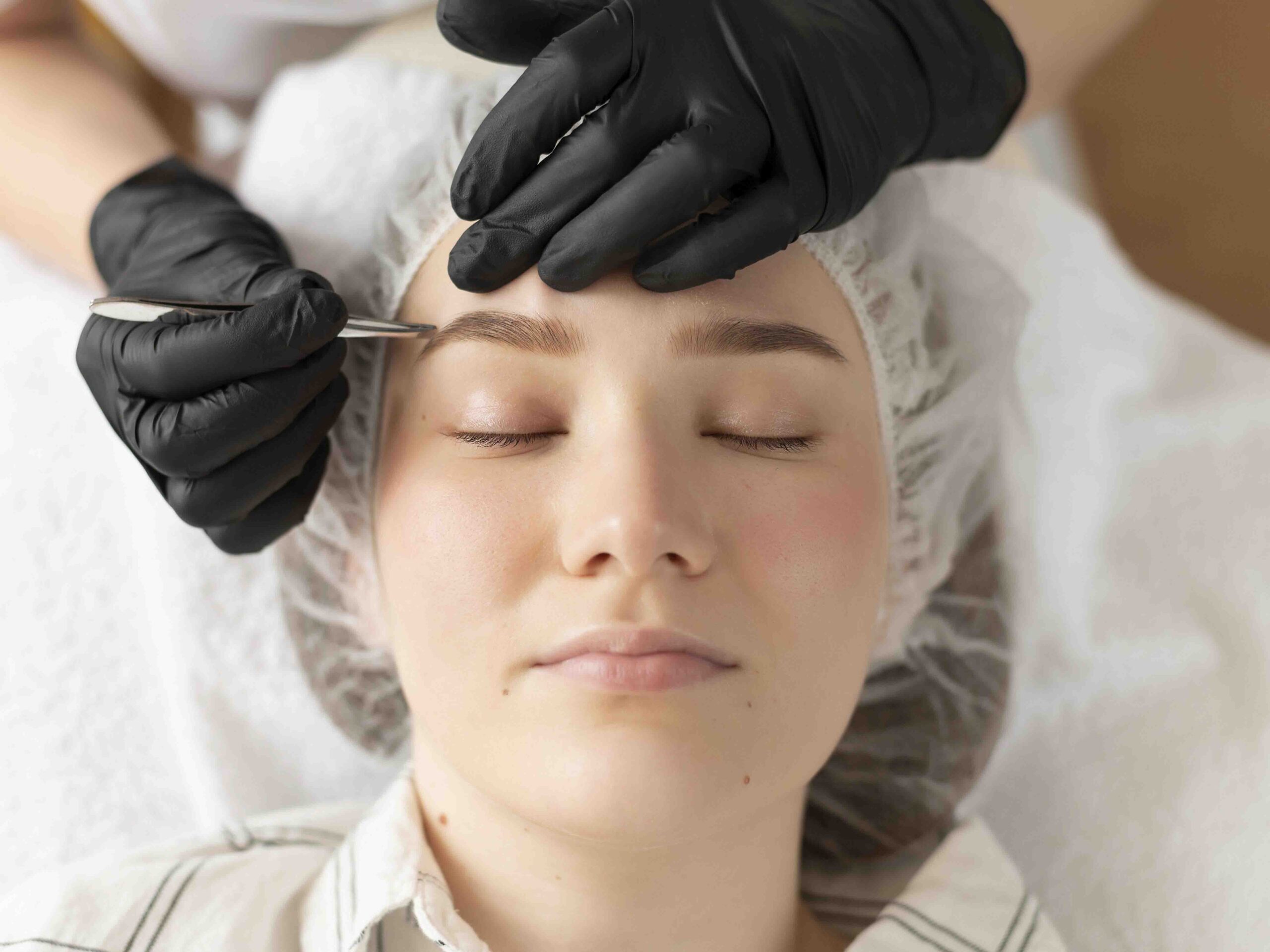 Woman receiving precise eyebrow hair restoration treatment from a specialist wearing black gloves.